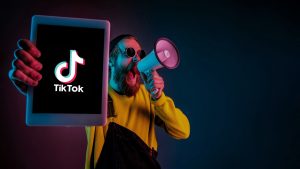 a man in a yellow jersey and on one hand he is holding a megaphone announcing how to increase TikTok views and go viral and on the other hand showing the screen of a tablet that shows the TikTok logo.