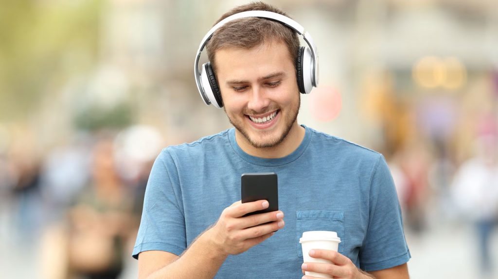 a happy man smiling and wearing headphones and watching TikTok videos on his phone with a coffee in his hand