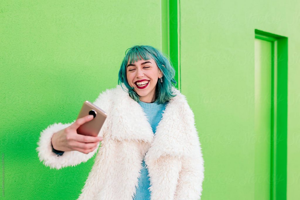 Happy young woman with green hair wearing a white coat and taking a selfie with her mobile phone.