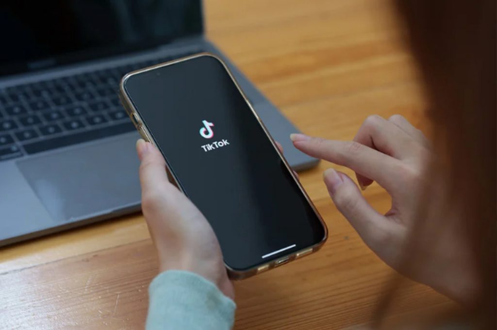 a smartphone in hands of a person who is touching the screen with her index finger and the screen of the hone showing the TikTok logo. In the background you can see a laptop on a desk.
