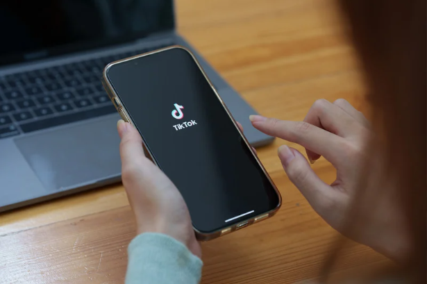 Hands holding an iPhone showing the TikTok app logo in the background there is a wooden desk that shows a mac laptop on it.