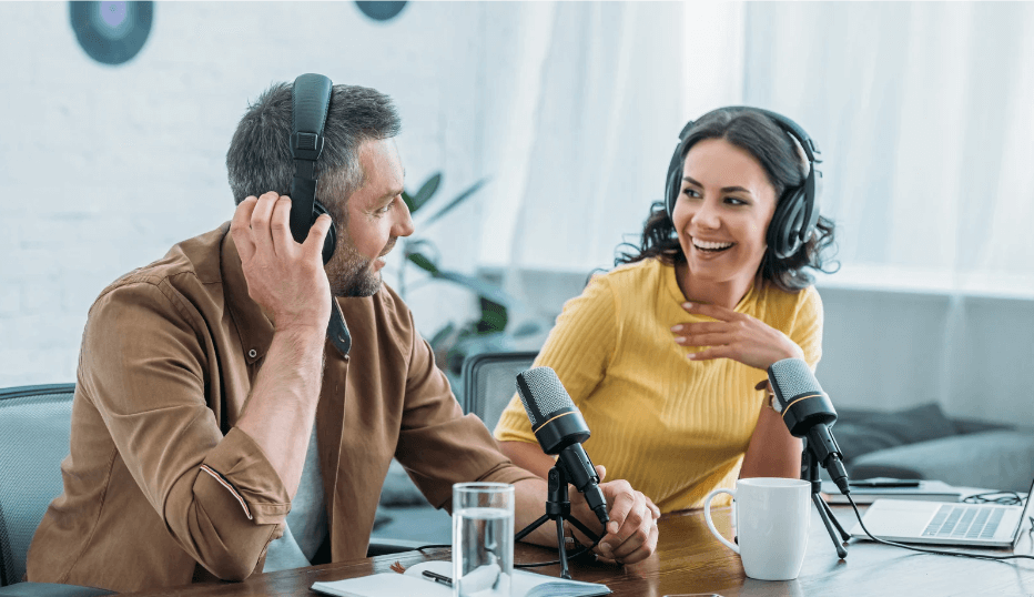 a man and woman sitting next to each other recording a Spotify podcast with headphones and vintage microphones.