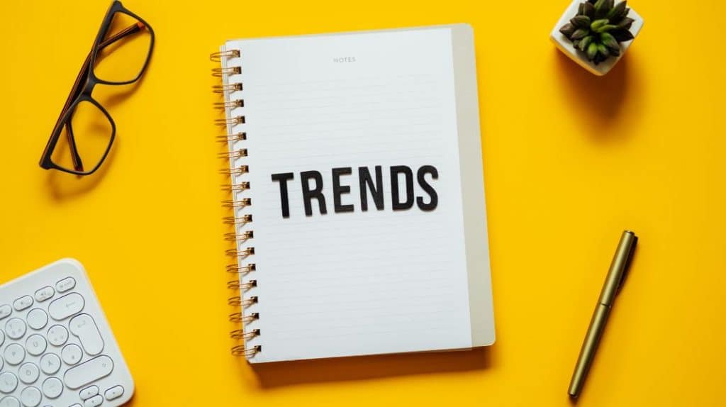 A picture of a notebook with the word Trends on it, glasses, a pen, a keyboard and a cactus
